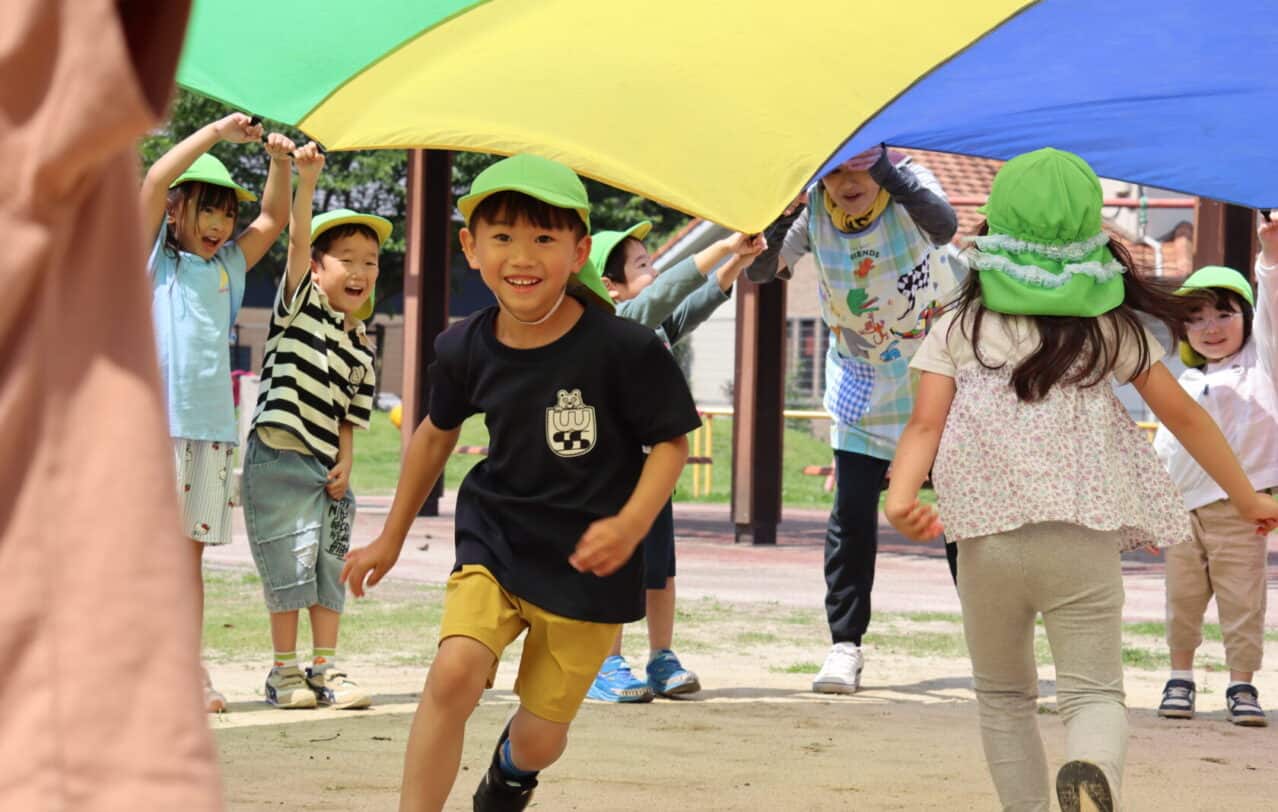熊本のヒルトップインターナショナルスクールで、カラフルなパラシュートの下を走る子供。A young child running underneath a colorful parachute at Hilltop International School in Kumamoto.
